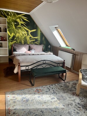 Un dormitorio con techo inclinado, una cama con un banco tapizado en verde, un radiador, una estantería y un ventanal grande.