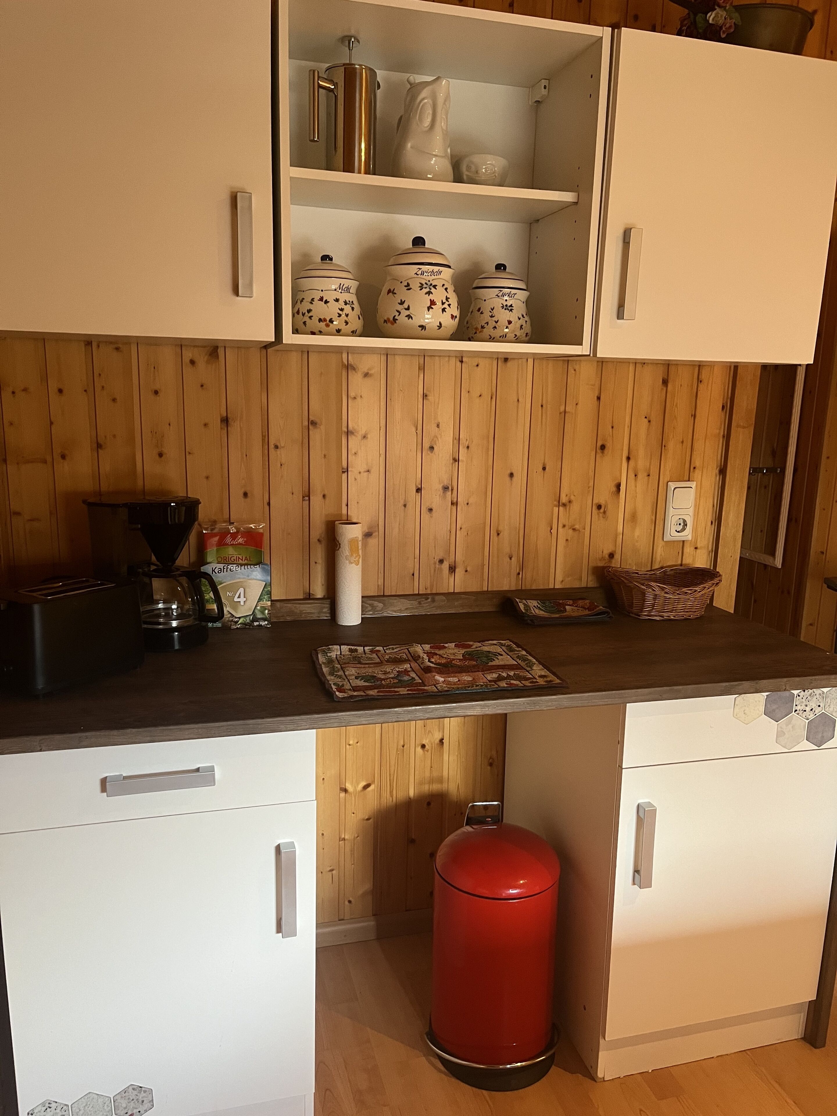 A kitchen with wooden paneling, a black countertop, and white cabinets. There are various kitchen items on the shelves, including a coffee maker, a kettle, and a basket.