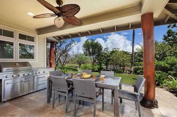 An outdoor kitchen with a grill, dining table, and chairs, surrounded by a garden and a view of the sky.