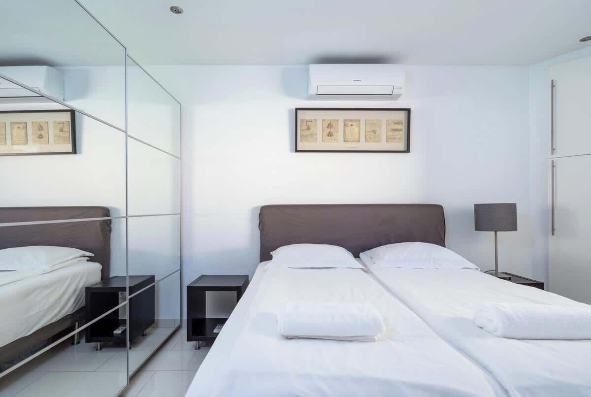 A modern hotel room with a large mirror, two beds, a bedside table, a lamp, and framed artwork on the wall.