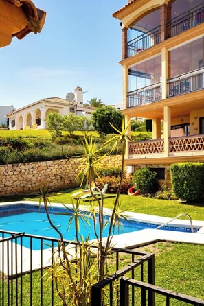 A pool area with a stone wall, a two-story building with balconies, and a white building with arches in the background.