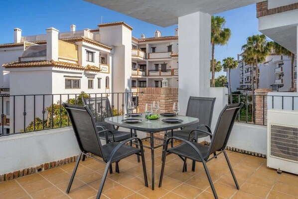 Een balkon met een ronde tafel en vier stoelen, met uitzicht op een woongebied met meerdere gebouwen en palmbomen.