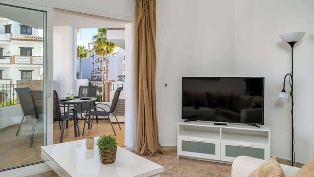 Een moderne woonkamer met een flatscreen televisie, een eethoek met stoelen en een tafel, en een balkon met uitzicht op andere gebouwen en palmbomen.