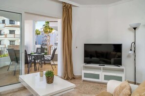 Een moderne woonkamer met een flatscreen televisie, een eethoek met stoelen en een tafel, en een balkon met uitzicht op andere gebouwen en palmbomen.
