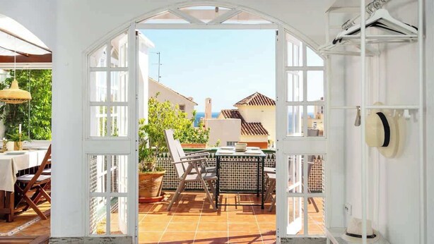 Un espace patio avec une table et des chaises, une porte blanche donnant sur l’extérieur et un portant à vêtements avec des chapeaux.