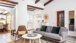 Un salon spacieux avec un canapé gris, une table basse ronde blanche et un fauteuil en osier. Le plafond est en bois avec des poutres apparentes, un mur blanc orné d’un grand miroir rond décoratif et une porte en bois avec des panneaux de verre.