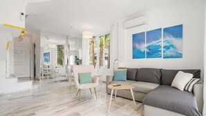 Un salon moderne avec un canapé gris, une petite table ronde et un fauteuil blanc. Il y a trois grandes fenêtres avec des rideaux, un coin repas à l’arrière-plan et une porte blanche.
