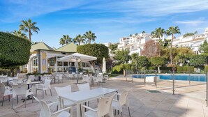 Une terrasse extérieure avec des tables et des chaises blanches, une piscine et un bâtiment doté d’un toit végétalisé en arrière-plan.