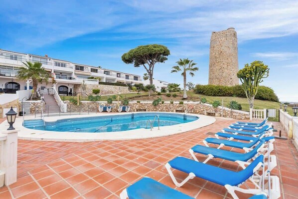 A swimming pool with blue lounge chairs, a stone tower, and a building with a staircase. - Charming seaview apt by Costarentals - Ref 49 (Las Lagunas de Mijas)