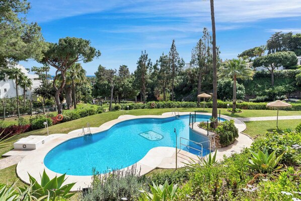 A swimming pool surrounded by greenery, a pathway, and a building in the background.