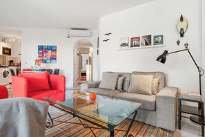 A modern living room with a red sofa, grey sofa, and a glass coffee table. There is a kitchen in the background, a washing machine, and a colorful painting on the wall.