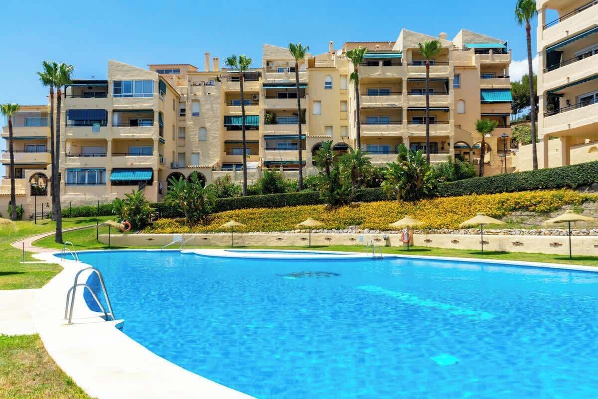 A swimming pool surrounded by a multi-story residential building with balconies, palm trees, and a landscaped area with flowers.