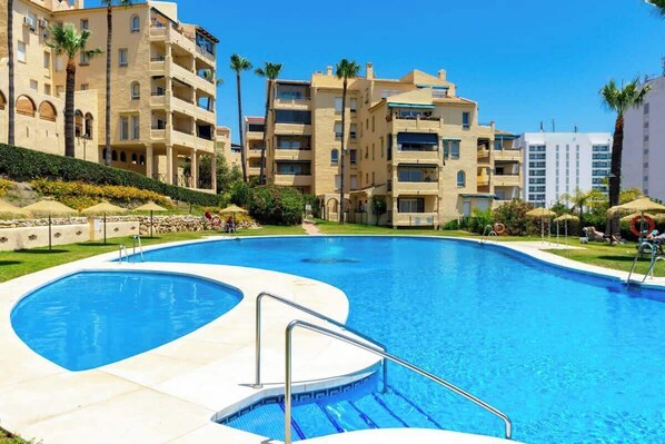 A swimming pool surrounded by multi-story buildings with balconies, palm trees, and a clear blue sky.