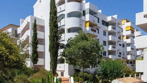 Un edificio blanco de varios pisos con balcones, rodeado de follaje y cactus.