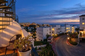 A coastal cityscape at dusk with modern buildings, a curved road, and a potted plant on a balcony.