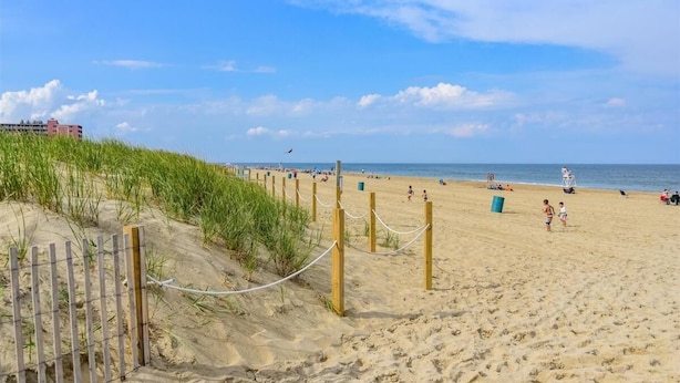 A sandy beach with a wooden fence and rope barrier, a clear sky, and a few people in the distance. - Beach view condo with private deck (Ocean City)