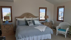 Ein Schlafzimmer mit einem großen Bett, zwei Nachttischen, einem Sessel und zwei Fenstern.