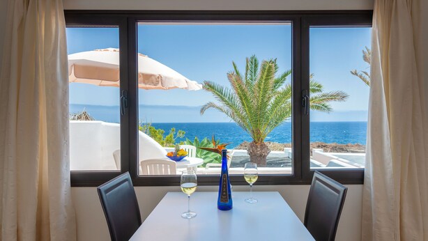 Una mesa con dos copas de vino y una botella, una palmera y vista al mar a través de una ventana.