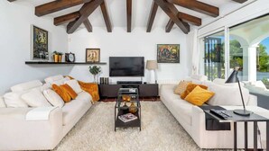 A modern living room with a high ceiling, wooden beams, a flat-screen TV, two sofas with cushions, a coffee table, and a large window offering a view. - Urb Villa with pool by Costarentals- Ref 139 (Mijas)