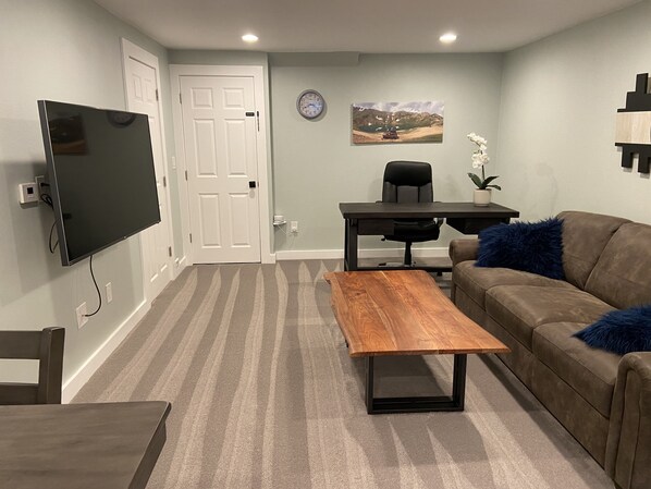 A living room with a flat-screen TV, a wooden coffee table, a brown sofa with a blue pillow, a desk with a chair, and a wall clock.