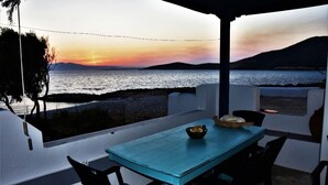 Een overdekte eethoek met een tafel en stoelen, met uitzicht op water en heuvels tijdens zonsondergang.