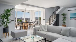 Een moderne woonkamer met een grijze bank, een glazen salontafel en een eethoek met tafel en stoelen. Er is een trap die naar een verdieping leidt, en grote ramen met uitzicht op de stad.