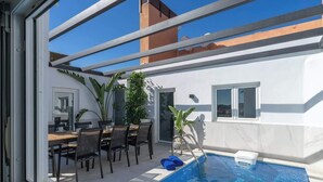 Een modern zwembadterras met een eethoek en een klein tafeltje, omgeven door potplanten.