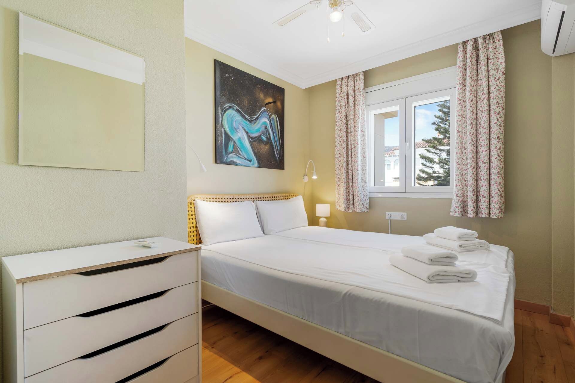 Quarto de hotel com cama, mesa de cabeceira, luminária, pintura e janela com cortinas.
