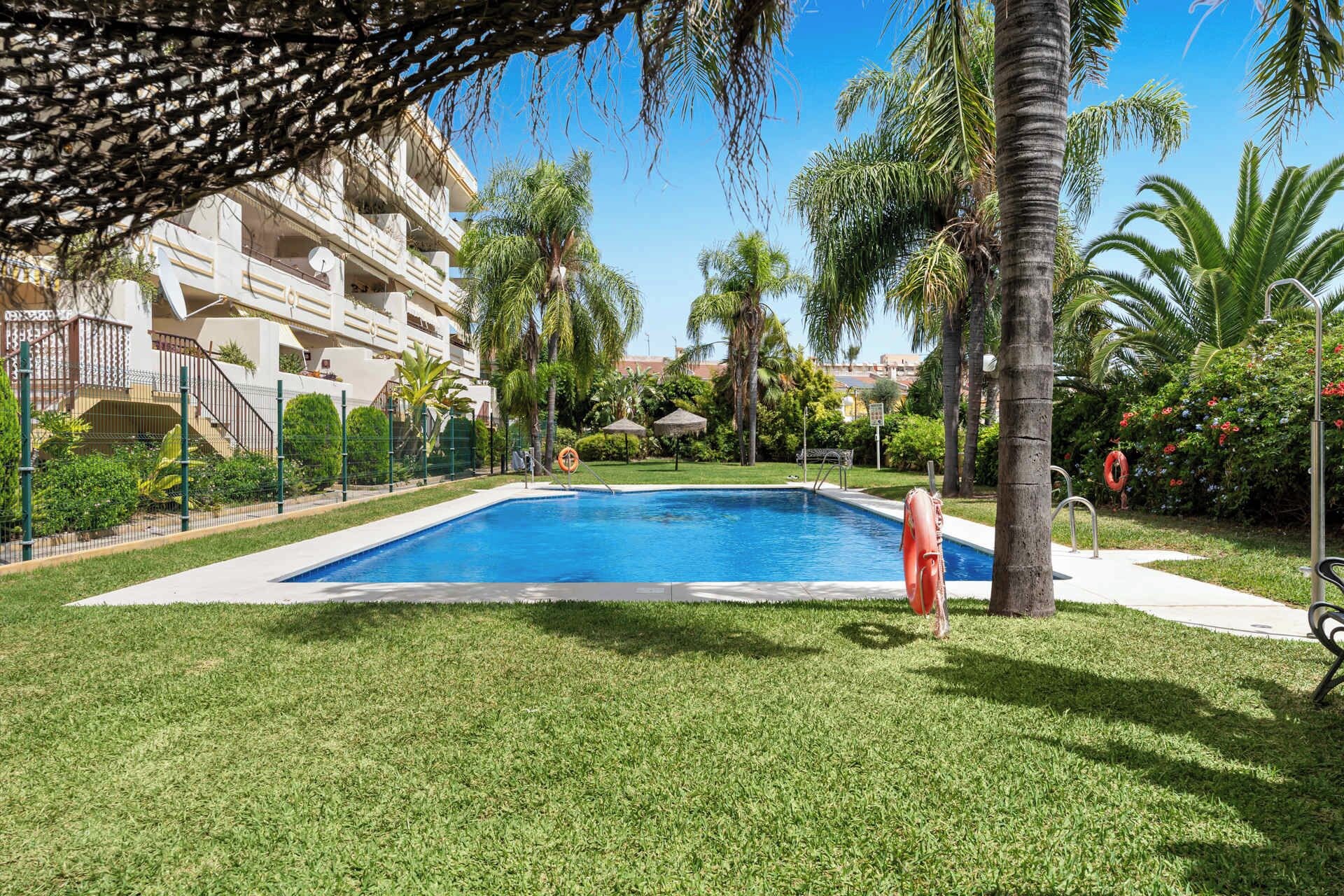 Une piscine entourée de pelouse bien entretenue, de palmiers et d’un immeuble à plusieurs étages doté de balcons.