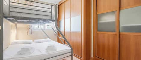 A bunk bed with a mattress, pillows, and folded towels. There is a small wooden table with a remote control. The room has wooden flooring and a wooden wardrobe with glass doors.