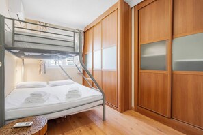 Un lit superposé avec un matelas, des oreillers et des serviettes pliées. Il y a une petite table en bois avec une télécommande. La pièce est dotée d’un plancher en bois et d’une armoire en bois avec des portes en verre.