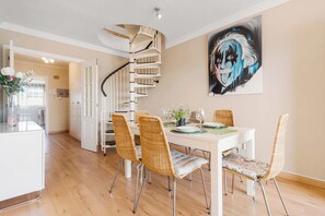 Un coin repas avec une table blanche, des chaises en osier, un escalier en colimaçon et un grand tableau abstrait accroché au mur.