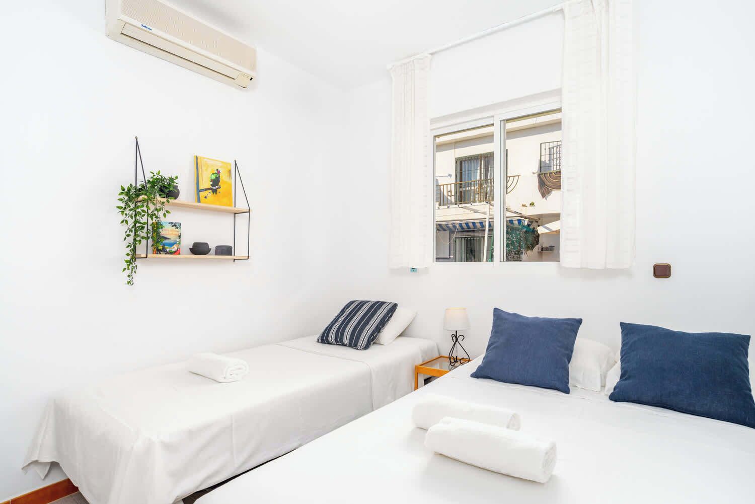 Een hotelkamer met twee bedden, een raam met gordijnen, een wandairconditioner, een plank met decoratieve objecten en een tafeltje met een lamp.