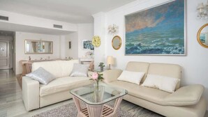 Un salon avec un canapé beige, une table basse en verre et un grand tableau représentant la mer.