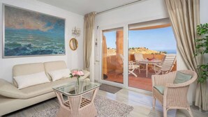 Un salon comprenant un canapé, une table basse en verre, des fauteuils en osier, un tableau représentant la mer et une porte-fenêtre coulissante donnant sur un espace extérieur aménagé pour se détendre.