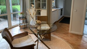 Ein Essbereich mit einem Glastisch, Korbstühlen und Blick auf einen Garten durch große Fenster.