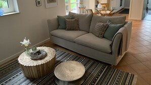 Ein Wohnzimmer mit grauem Sofa, einem runden Holztisch und einem Streifen Teppich. Es gibt einen Essbereich mit Glastisch und Stühlen, und im Hintergrund ist eine Küche zu sehen.