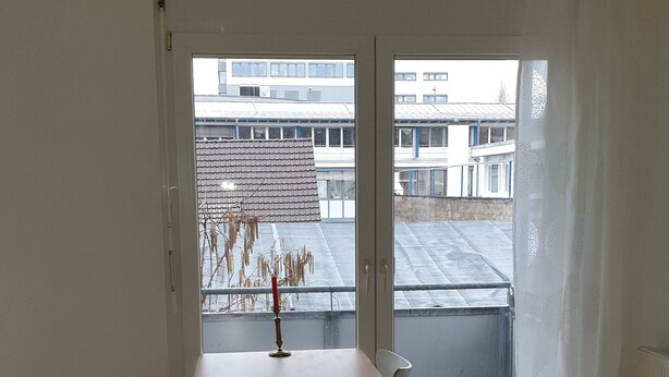 Ein Zimmer mit Holzboden, einem weißen Tisch und einem Stuhl. Ein Fenster bietet Blick auf Gebäude außerhalb.