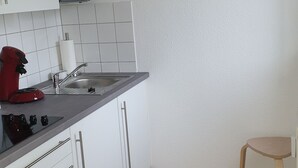 Eine Küche mit weißen Schränken, einer Mikrowelle, einem Spülbecken, einem roten Wasserkocher und einem Holzstuhl.