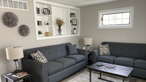 A modern living room with two grey sofas, a coffee table, and a side table with a lamp. There is a white shelving unit with decorative items and a window above the sofas. - In the Heart of Three Oaks - Steps to Downtown (Three Oaks)