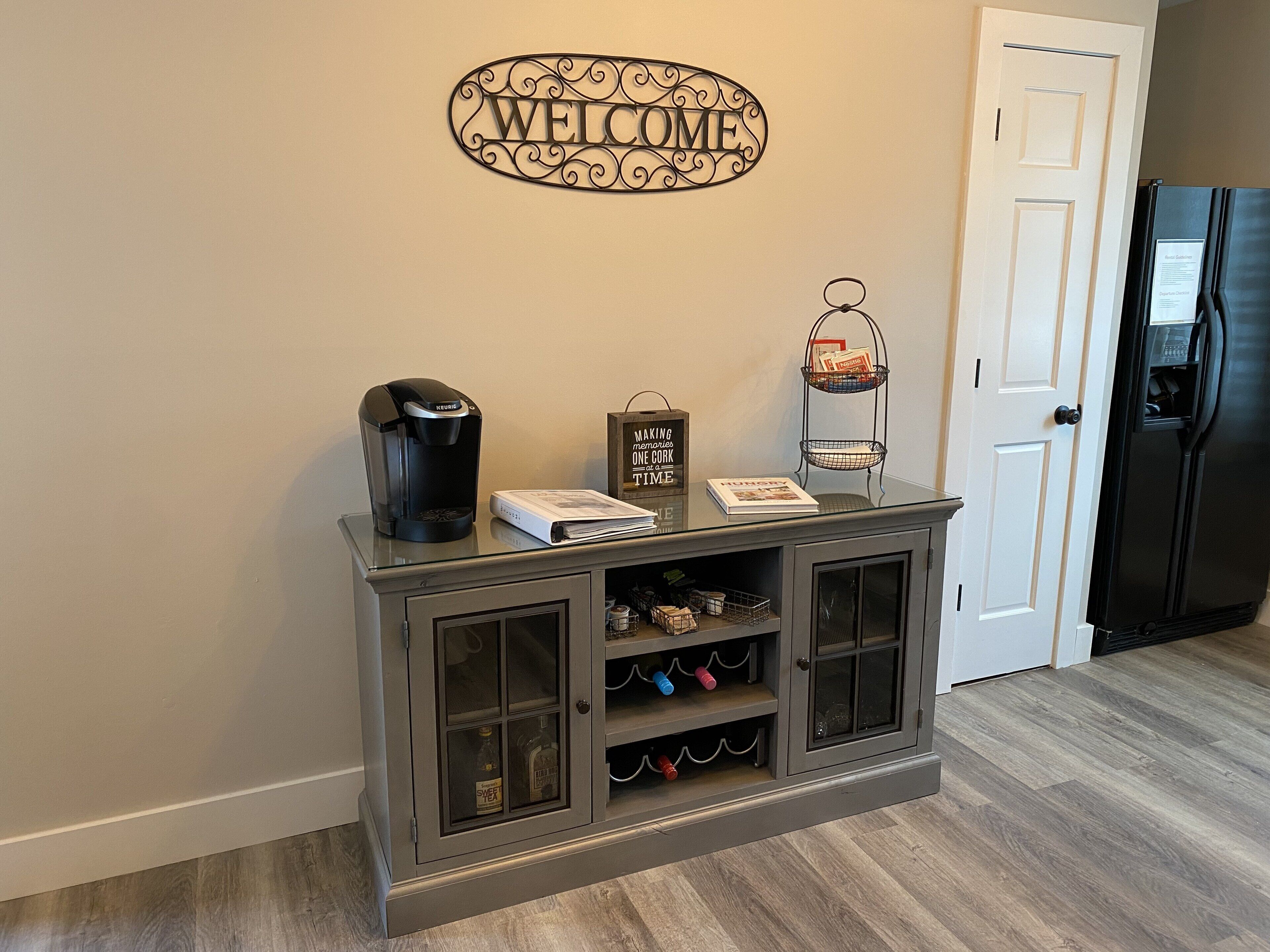 A kitchen with a coffee maker, a welcome sign, a small book, and a wine rack.