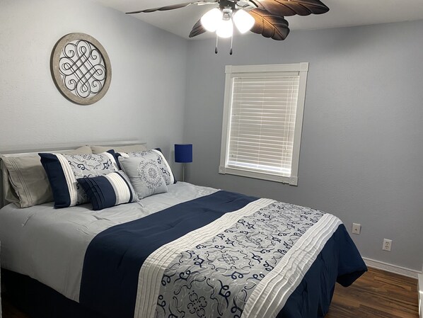 Une chambre à coucher comprenant un lit, un ventilateur de plafond, une fenêtre avec des stores et un luminaire fixé au mur.
