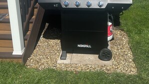 An outdoor NEXGRILL barbecue grill with four burners, positioned on a deck with a wooden railing and a white house in the background. - In the Heart of Three Oaks - Steps to Downtown (Three Oaks)