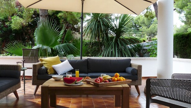 Un área de estar al aire libre con un sofá acolchado, una mesa de madera con frutas y bebidas, y un gran paraguas que proporciona sombra.