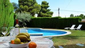 Escena junto a la piscina con una mesa que sostiene una botella, vasos y fruta.
