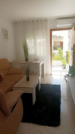 A living room with a sofa, a coffee table, and a plant. There is a window with curtains and a view of the outside. - A beautiful bungalow (Torrevieja)