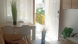 Een woonkamer met een bank, een bijzettafeltje en een plant in een pot. Er is een deur die leidt naar een buitenruimte met een geel hek.