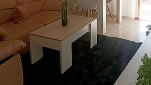 Een woonkamer met een bank, een salontafel en een eettafel met stoelen. Er staan twee potplanten, een op de salontafel en een op de eettafel. De kamer heeft een zwart vloerkleed en witte gordijnen.