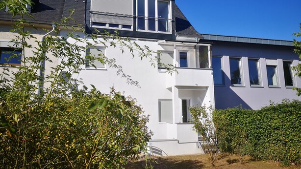 Une maison à deux étages, à l’extérieur blanc, dotée d’un balcon avec une rambarde en métal et d’un petit jardin agrémenté d’arbustes et d’un arbre.