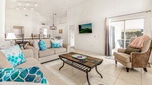 A spacious living room with a sofa, armchair, and coffee table. There is a television mounted on the wall and a ceiling fan.